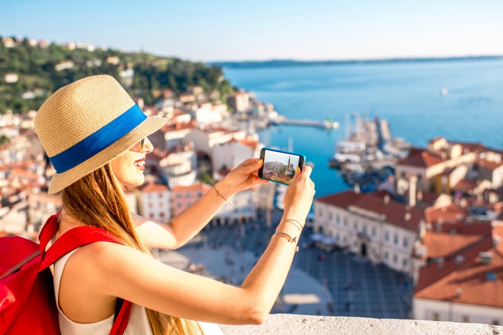 Woman in hat takes a photo of seaside town with smartphone, overlooking harbor and rooftops.
