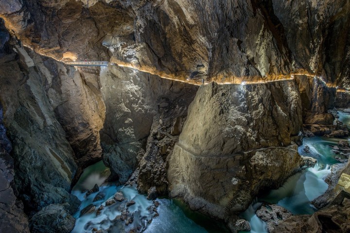 A narrow walkway with lights inside a large rocky cave above a flowing river.