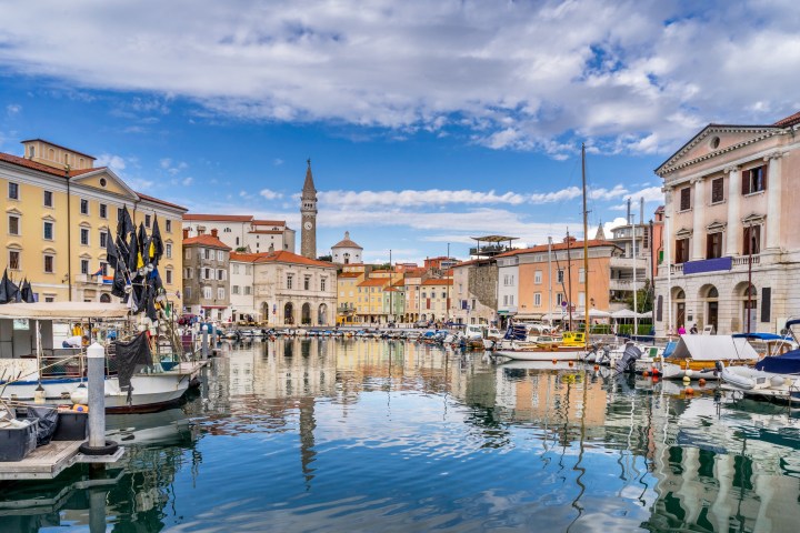 Picturesque harbor with boats, colorful buildings, and a bell tower under a blue sky with clouds.