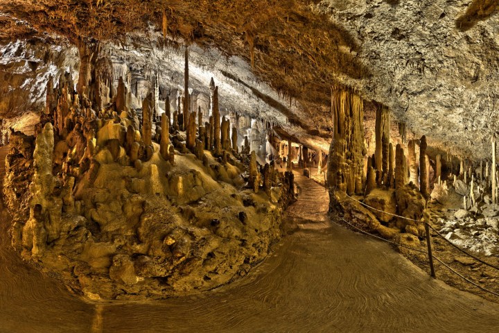 Cave interior with stalagmites, stalactites, and illuminated pathway.