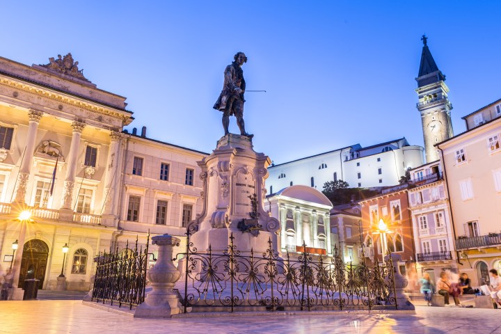 Illuminated square with a historic statue, buildings, and clock tower at dusk.