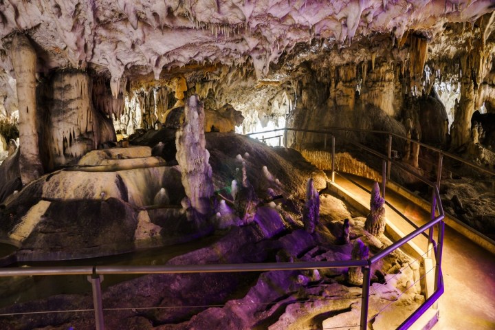 Illuminated cave interior with stalactites, stalagmites, and a walkway.