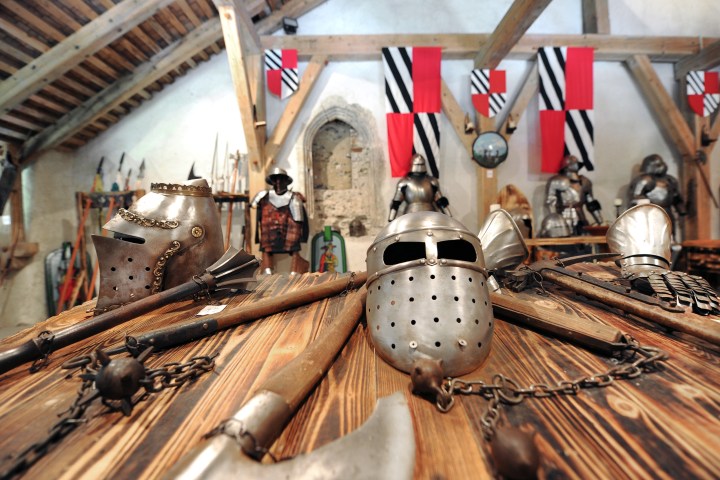 Medieval helmets and weapons displayed on a wooden table in a rustic room with armor and banners.