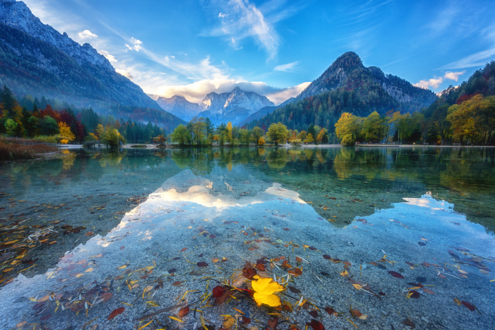 Mountain lake with autumn leaves, reflections, and misty peaks under a blue sky.