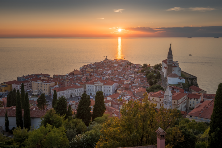 Sunset over coastal town with red roofs, church tower, and sea in the background.