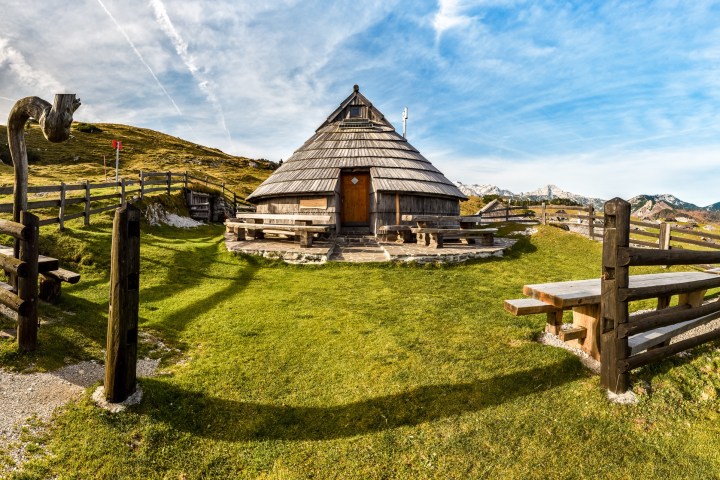 Wooden hut with conical roof surrounded by fence on grassy hill under blue sky.