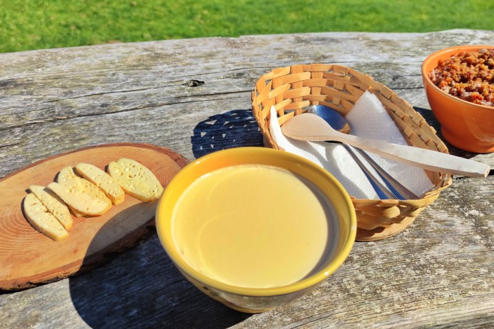 Outdoor table with sliced cheese, yellow soup in bowl, utensils, and a bowl with crumbly mixture.