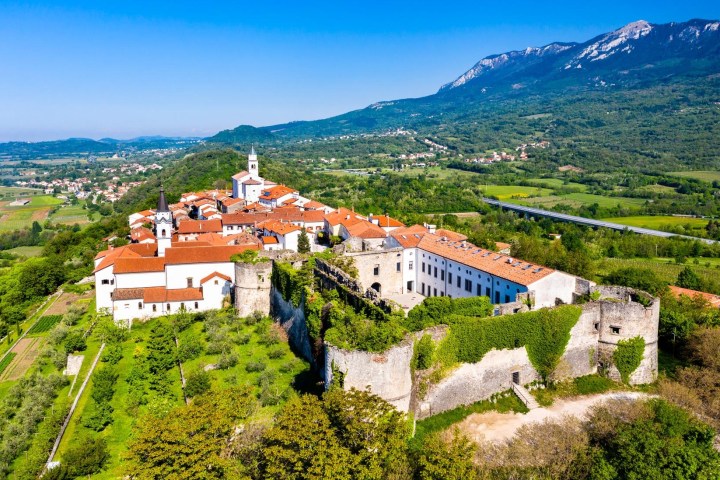 Aerial view of a hilltop castle with red roofs and greenery, set against mountains and a blue sky.