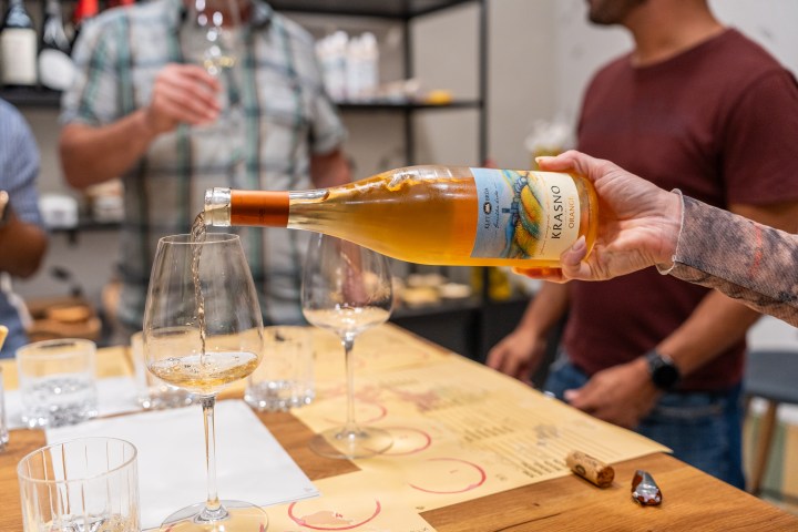 Person pouring wine into a glass at a tasting, with people in the background.
