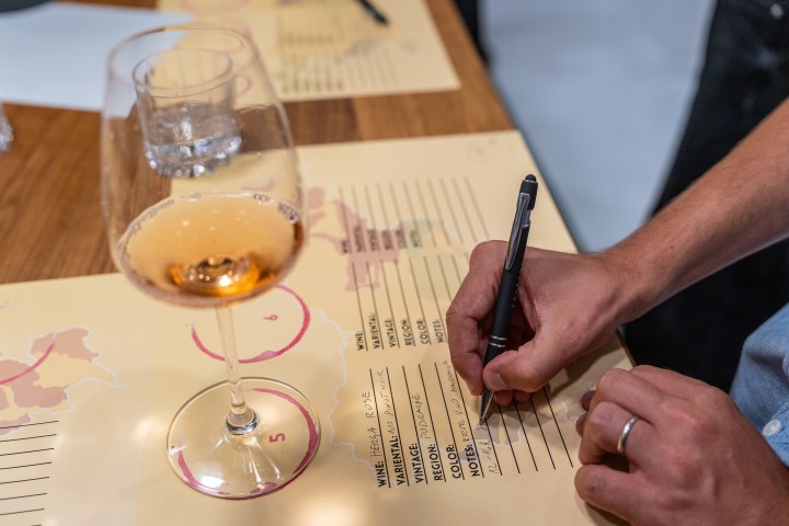 Person writing on wine tasting sheet next to a glass of rosé wine on a wooden table.