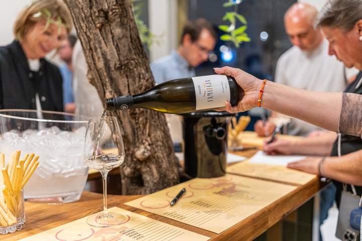 Person pouring white wine into a glass at a tasting event with people in the background.