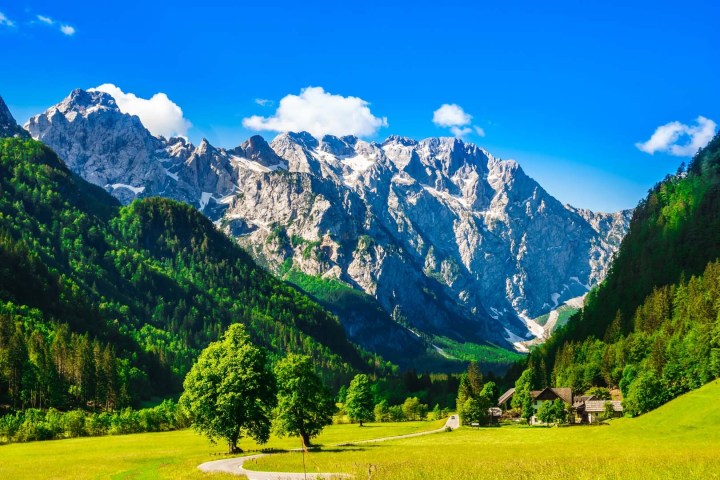 Mountain landscape with green valley, trees, and buildings under a blue sky with clouds.