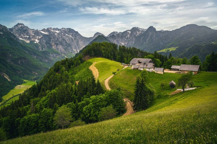 Scenic mountain view with lush green hills, trees, and rustic farm buildings.