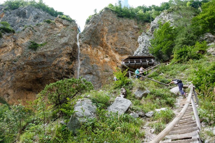 People climbing stairs to a viewing platform near a mountain waterfall, surrounded by greenery.