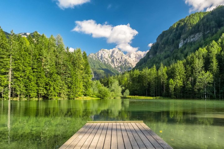 Wooden pier extending into a tranquil lake surrounded by lush green forest and distant mountains.